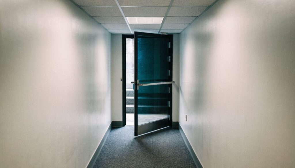 Porte de sortie de secours entrouverte équipée d'une barre anti-panique horizontale, au bout d'un couloir