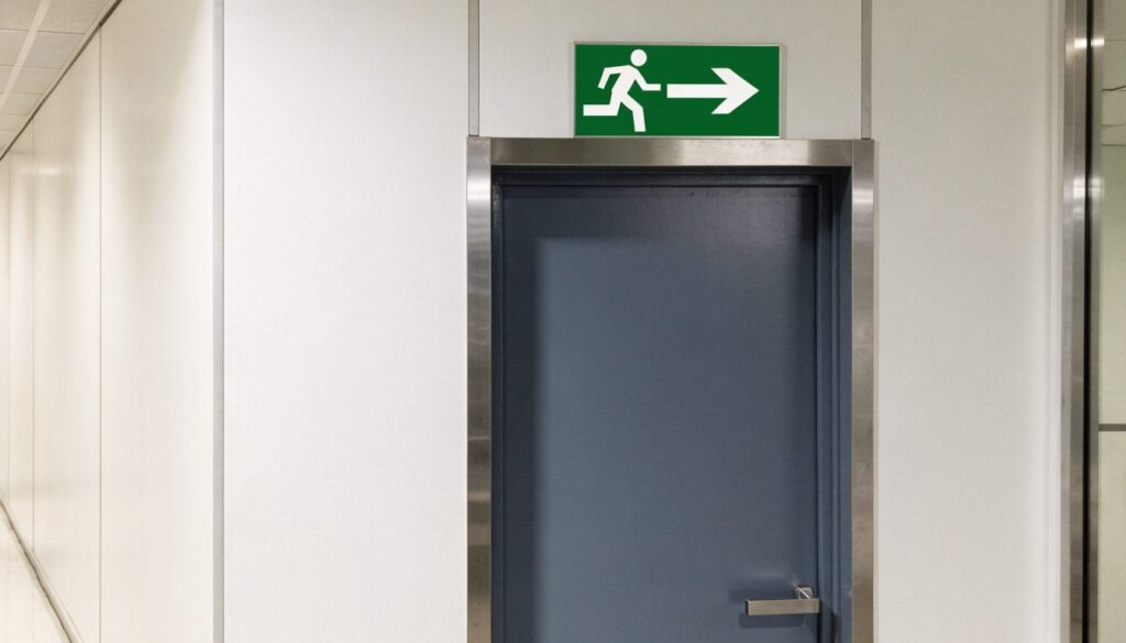 Panneau rectangulaire vert d'issue de secours montrant une silhouette qui court et une flèche, fixé au-dessus d'une porte de couloir